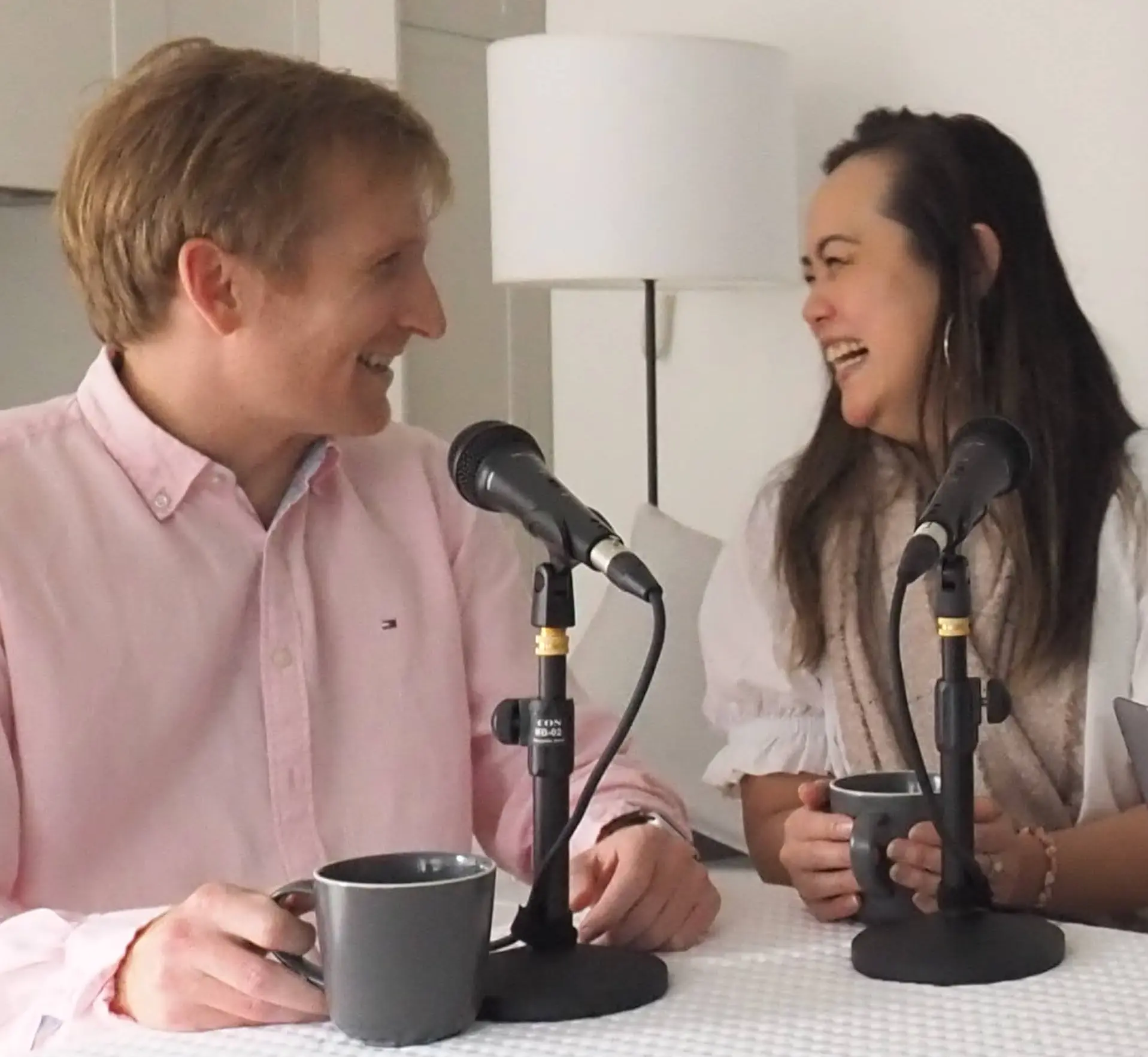 A man and a woman sitting behind microphones, looking at each other smiling
